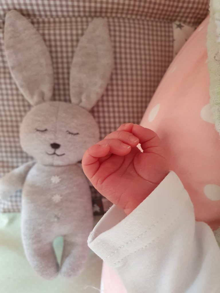 Ein Bild, das einen Baby-Arm zeigt. Im Hintergrund liegt ein Kuscheltier, ein grauer Hase.