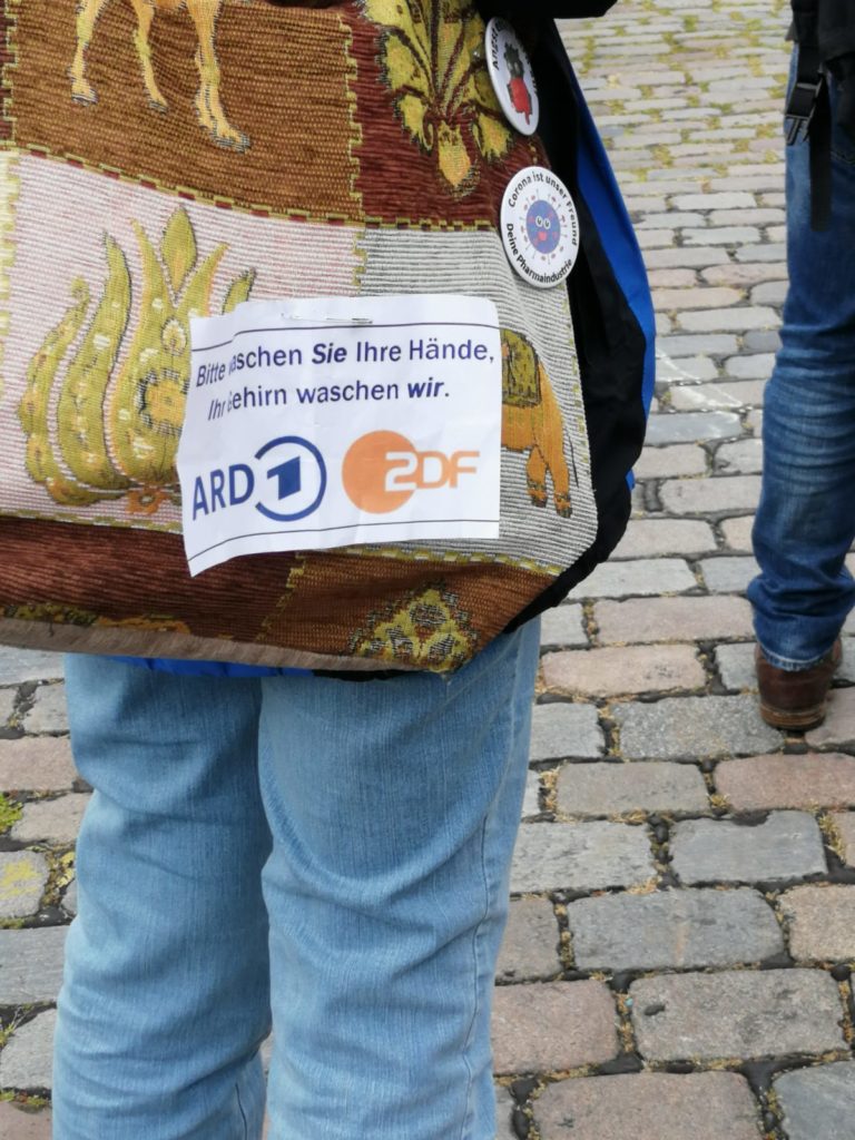 Ein Zettel an der Tasche einer Demonstrantin, auf dem steht: "Bitte waschen Sie Ihre Hände. Ihr Gehirn waschen wir.", gefolgt von den Logos von ARD und ZDF.